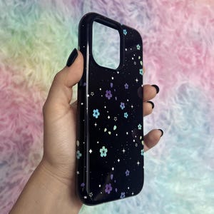 May include: Black phone case with a floral pattern of small purple, blue, and green flowers on a black background with white stars.