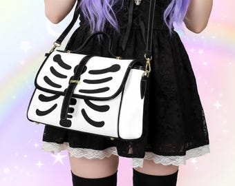 以下が含まれることがあります： 黒い骸骨の肋骨のデザインが施された白いハンドバッグ。バッグは、黒いストラップと金色の金具が付いたフラップ開閉式です。バッグにはトップハンドルとショルダーストラップが付いています。ユニークなアクセサリーです。