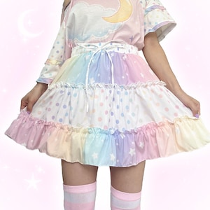 May include: A tiered skirt featuring a pastel rainbow colour palette with white star patterns on the bottom ruffle and a white polka dot print on the middle section. The skirt has a white drawstring waist and a ruffled hemline with a flared silhouette.