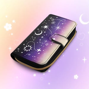 May include: A black and purple wallet with a celestial design featuring stars, moons, and suns. The wallet has a gold zipper and a snap closure. The design fades from black to purple to white.