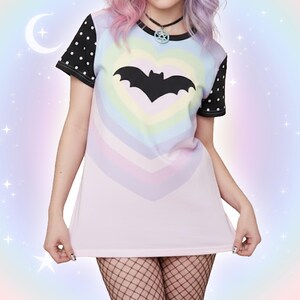May include: A pastel-colored t-shirt featuring a black bat inside a rainbow heart design. The shirt has black polka dot sleeves and a black collar. The overall aesthetic is a blend of goth and pastel styles, with a soft, dreamy background.