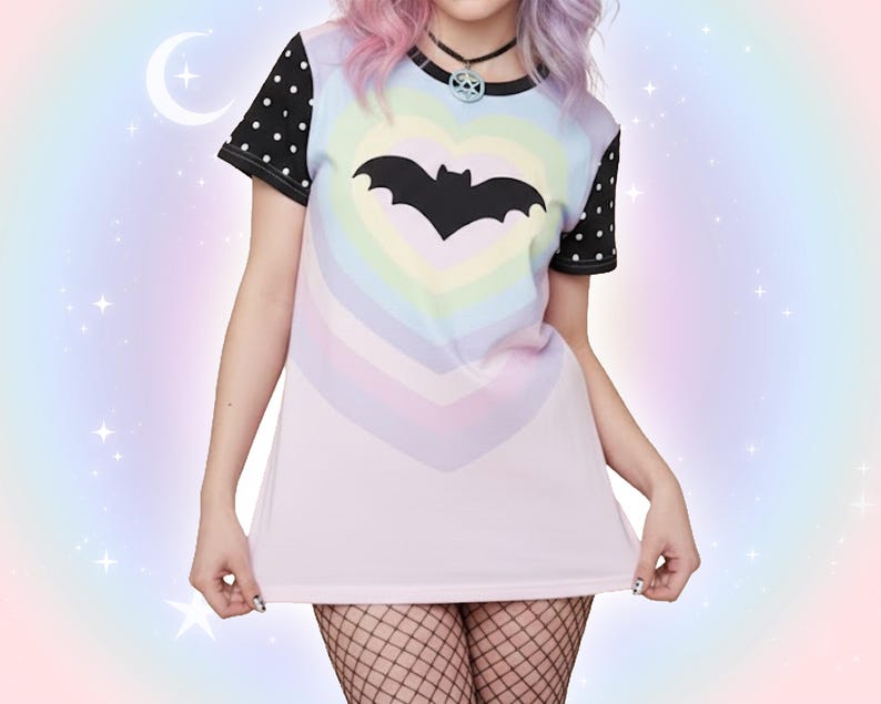 May include: A pastel-colored t-shirt featuring a black bat inside a rainbow heart design. The shirt has black polka dot sleeves and a black collar. The overall aesthetic is a blend of goth and pastel styles, with a soft, dreamy background.