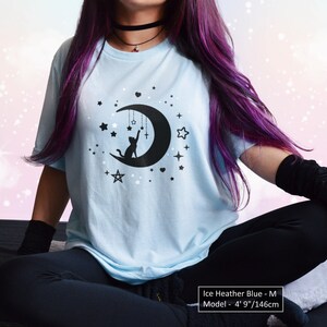 May include: Ice heather blue t-shirt featuring a black graphic of a cat reaching for stars and a crescent moon. The shirt has a crew neck and short sleeves. The model is 4' 9" tall.