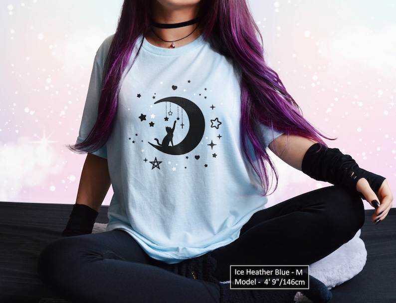 May include: Ice heather blue t-shirt featuring a black graphic of a cat reaching for stars and a crescent moon. The shirt has a crew neck and short sleeves. The model is 4' 9" tall.
