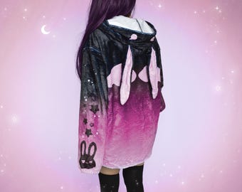 Könnte beinhalten: Ein schwarzer und rosa Ombre-Hoodie mit einem großen rosa Hasen-Design auf der Rückseite. Der Hoodie hat eine schwarze Kapuze und ein weißes Futter. Der Hoodie ist mit kleinen schwarzen Sternen verziert.