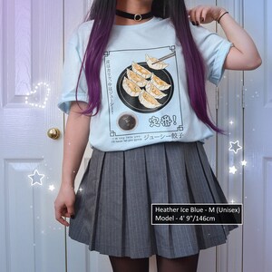 May include: A light blue t-shirt with a graphic of dumplings and the text "皮はカリッ、中はジューシー! 定番! ジューシー餃子" in Japanese. The model is wearing a grey pleated skirt and has purple hair. The text "Heather Ice Blue - M (Unisex) Model - 4' 9"/146cm" is at the bottom of the image.