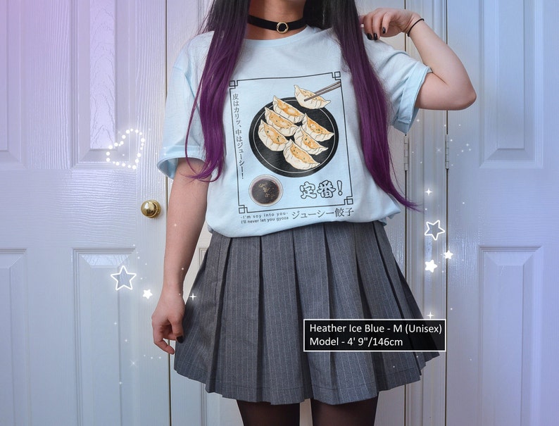 May include: A light blue t-shirt with a graphic of dumplings and the text "皮はカリッ、中はジューシー! 定番! ジューシー餃子" in Japanese. The model is wearing a grey pleated skirt and has purple hair. The text "Heather Ice Blue - M (Unisex) Model - 4' 9"/146cm" is at the bottom of the image.