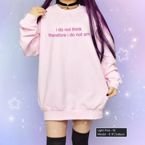 May include: Light pink sweatshirt with the text "i do not think therefore i do not am" in a soft, rounded font. The model is wearing a black choker. The background is a gradient of purple and blue with white star accents.