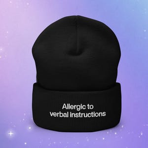 Op de afbeelding: Zwarte beanie met witte tekst die "Allergie voor verbale instructies" zegt.