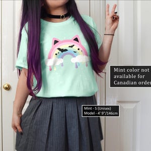 K&ouml;nnte beinhalten: Ein mintgr&uuml;nes T-Shirt mit einem bunten, katzenf&ouml;rmigen Regenbogen-Design mit Flederm&auml;usen und tropfenden Wolken. Das Shirt ist in der Unisex-Gr&ouml;&szlig;e S. Das Model ist 146 cm gro&szlig;. Der Text auf dem Shirt lautet "Mint color not available for Canadian orders."