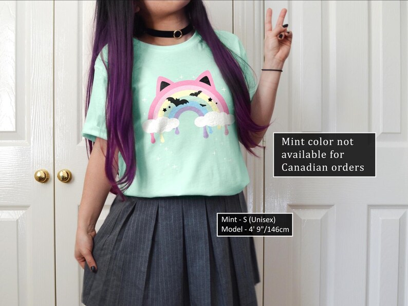 K&ouml;nnte beinhalten: Ein mintgr&uuml;nes T-Shirt mit einem bunten, katzenf&ouml;rmigen Regenbogen-Design mit Flederm&auml;usen und tropfenden Wolken. Das Shirt ist in der Unisex-Gr&ouml;&szlig;e S. Das Model ist 146 cm gro&szlig;. Der Text auf dem Shirt lautet "Mint color not available for Canadian orders."