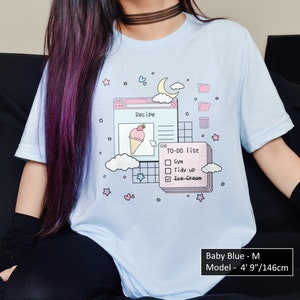 May include: A light blue t-shirt featuring a graphic of a computer screen with a recipe, a to-do list, and a cloud. The text on the screen reads "Recipe" and "TO-DO list". The to-do list includes "Gym", "Tidy up", and "Ice-Cream". The graphic is in a pastel colour scheme with pink, blue, and white. The t-shirt is available in size M.