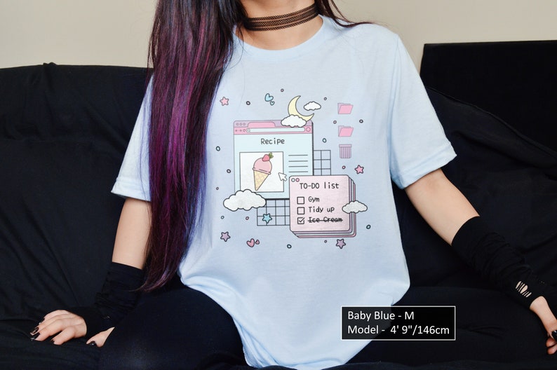 May include: A light blue t-shirt featuring a graphic of a computer screen with a recipe, a to-do list, and a cloud. The text on the screen reads "Recipe" and "TO-DO list". The to-do list includes "Gym", "Tidy up", and "Ice-Cream". The graphic is in a pastel colour scheme with pink, blue, and white. The t-shirt is available in size M.
