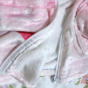 Puede incluir: Una chaqueta de forro polar rosa y blanca con cierre de cremallera. La chaqueta tiene un estampado de fresas en la parte delantera y trasera.