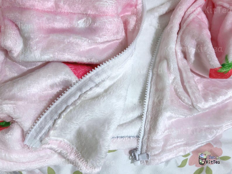 Puede incluir: Una chaqueta de forro polar rosa y blanca con cierre de cremallera. La chaqueta tiene un estampado de fresas en la parte delantera y trasera.