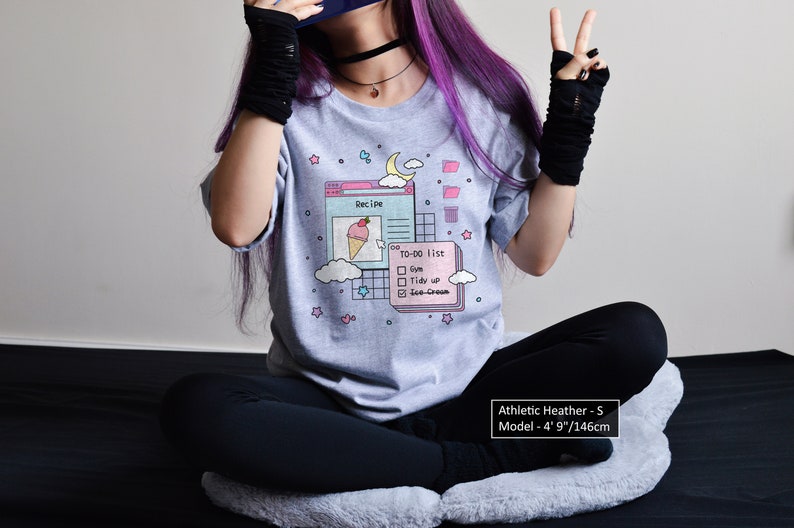 May include: A heather grey t-shirt featuring a graphic of a computer screen with a recipe and a to-do list. The graphic includes a pink ice cream cone, a blue cloud, and a yellow moon. The text on the shirt reads "Recipe" and "To-Do List". The model is wearing a size S.