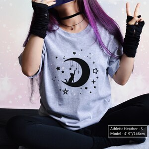 May include: Athletic heather gray t-shirt with a black graphic of a cat reaching for a star on a crescent moon, surrounded by stars. The text on the shirt says "Athletic Heather - S". The model is 4' 9"/146cm tall.