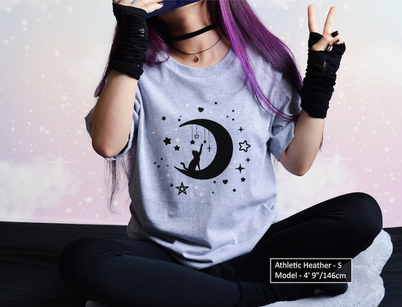 May include: Athletic heather gray t-shirt with a black graphic of a cat reaching for a star on a crescent moon, surrounded by stars. The text on the shirt says "Athletic Heather - S". The model is 4' 9"/146cm tall.