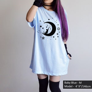 May include: Baby blue t-shirt with a black graphic of a cat sitting on a crescent moon, surrounded by stars. The shirt has short sleeves and a relaxed fit. The text "Baby Blue - M" and model measurements are visible.