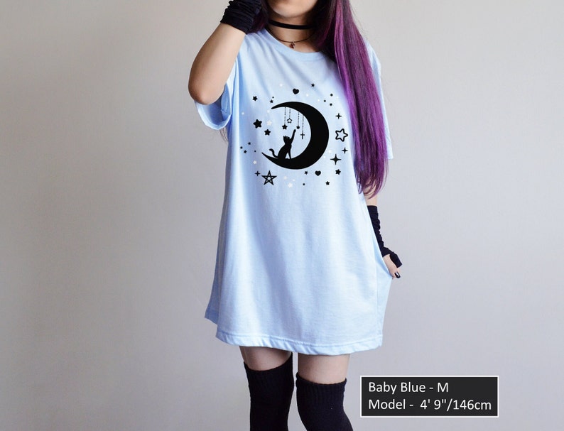 May include: Baby blue t-shirt with a black graphic of a cat sitting on a crescent moon, surrounded by stars. The shirt has short sleeves and a relaxed fit. The text "Baby Blue - M" and model measurements are visible.