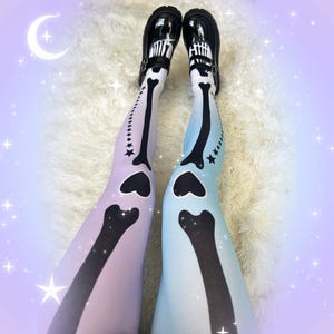 May include: Opaque tights featuring a gradient skeleton bone print in black against a pastel lavender and light blue background. The design includes heart-shaped knee joints and a row of small stars along the outer leg. Black patent leather shoes.