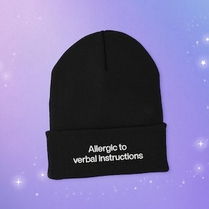 A cuffed beanie knit cap with a embroidered white text that says: A llergic to verbal instructions.