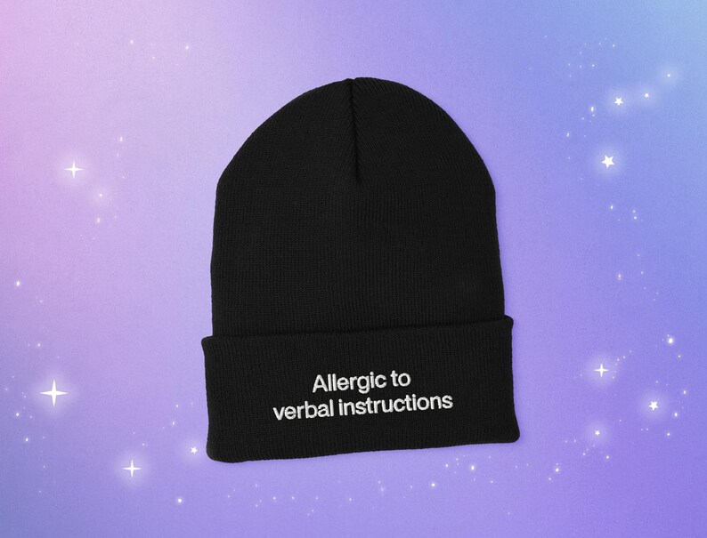 A cuffed beanie knit cap with a embroidered white text that says: A llergic to verbal instructions.