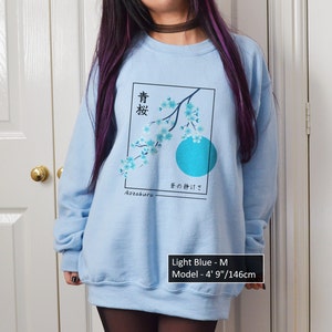 Pode incluir: Camisola azul claro com uma estampa gráfica de um ramo de cerejeira em flor e um círculo azul. O texto japonês na estampa diz "青桜 その静けさ Aozakura".