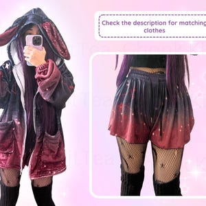 May include: A red and black hooded jacket with bunny ears and a galaxy print. The jacket is worn over a black shirt and black leggings. The person is holding a phone in front of their face. The image also shows a pair of red and black shorts with a galaxy print.