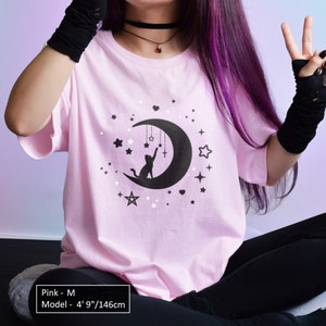 May include: A light pink t-shirt with a black graphic of a crescent moon, stars, and a cat reaching for a cross. The shirt is a crew neck style. The model is 4' 9"/146cm tall. The shirt is size M.