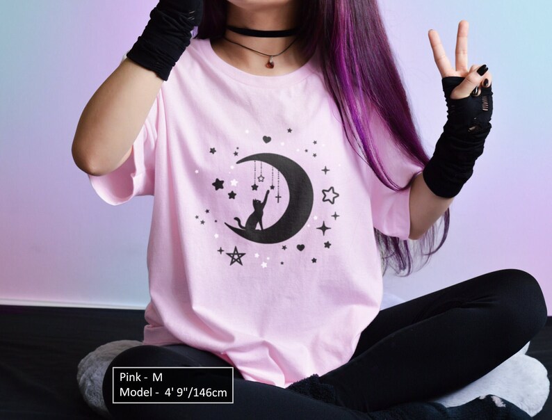 May include: A light pink t-shirt with a black graphic of a crescent moon, stars, and a cat reaching for a cross. The shirt is a crew neck style. The model is 4' 9"/146cm tall. The shirt is size M.