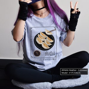 May include: A heather grey t-shirt with a graphic of a plate of dumplings and chopsticks. The text on the shirt reads "中度はカリッ、中はジューシー! 定番! ジューシー餃子". The model is 4' 9" tall.