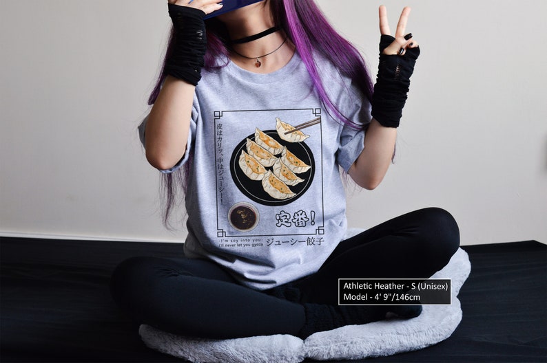 May include: A heather grey t-shirt with a graphic of a plate of dumplings and chopsticks. The text on the shirt reads "中度はカリッ、中はジューシー! 定番! ジューシー餃子". The model is 4' 9" tall.