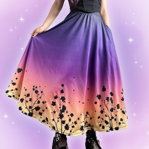 Puede incluir: Una falda larga y fluida con un degradado de morado, rosa y amarillo. La falda presenta un diseño floral negro en la parte inferior. La persona lleva zapatos negros y un top corsé negro.