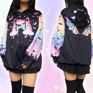May include: Black hoodie with a colourful rainbow, bats, and stars design. The sleeves have a pastel gradient. The hood features bat ears. The back of the hoodie has a bat wing design. The hoodie is worn with a black skirt.