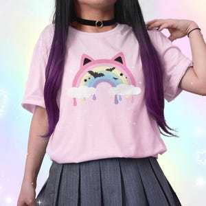 K&ouml;nnte beinhalten: Hellrosa T-Shirt mit einem Regenbogen-Design mit Katzenohren, Flederm&auml;usen und tropfenden Wolken. Das Shirt wird mit einem grauen Faltenrock getragen. Der Stil kombiniert niedliche und gruselige Elemente.