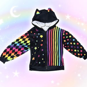 May include: Black zip-up hoodie with cat ears on the hood. The sleeves feature colorful patterns: one with rainbow diamonds, the other with polka dots. The front has a vertical rainbow stripe design and a star pattern.