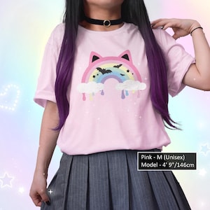 K&ouml;nnte beinhalten: Rosa Unisex-T-Shirt mit einem Regenbogen- und Fledermaus-Motiv. Der Regenbogen hat Katzenohren und befindet sich &uuml;ber Wolken mit tropfender Farbe. Das Model tr&auml;gt einen schwarzen Choker und einen grauen Faltenrock. Der Text sagt "Pink - M (Unisex) Model - 146cm".
