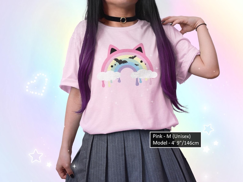 K&ouml;nnte beinhalten: Rosa Unisex-T-Shirt mit einem Regenbogen- und Fledermaus-Motiv. Der Regenbogen hat Katzenohren und befindet sich &uuml;ber Wolken mit tropfender Farbe. Das Model tr&auml;gt einen schwarzen Choker und einen grauen Faltenrock. Der Text sagt "Pink - M (Unisex) Model - 146cm".