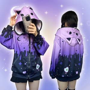 May include: A purple and black cat-themed hoodie with a zip-up front. The hood has a cat face design with ears, and a celestial pattern with stars, moons, and dripping paint details. The sleeves are black.