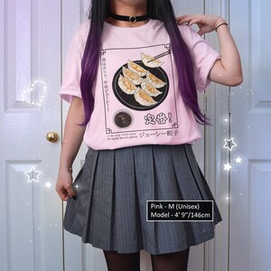May include: Pink t-shirt with a graphic of a plate of dumplings and chopsticks. The text on the shirt reads "皮はカリッ、中はジューシー! 定番! ジューシー餃子". The model is wearing a gray pleated skirt and is 4' 9" tall.