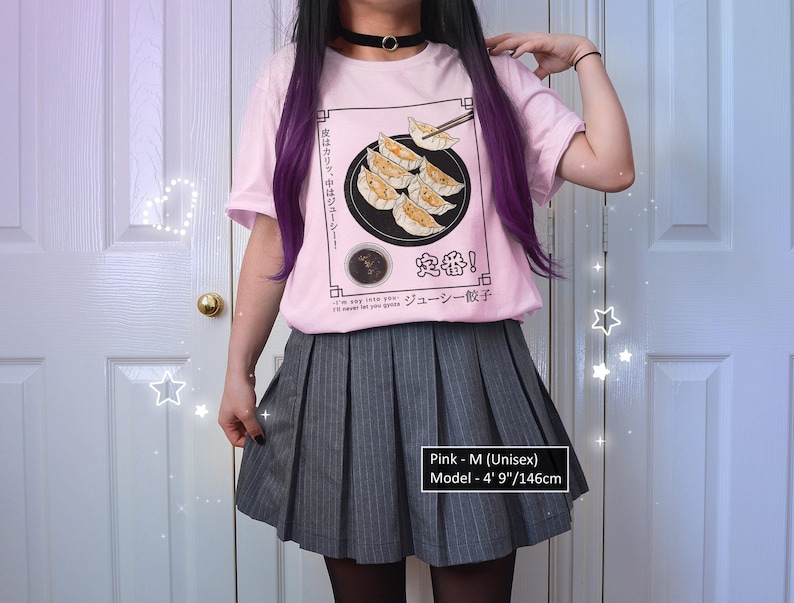 May include: Pink t-shirt with a graphic of a plate of dumplings and chopsticks. The text on the shirt reads "皮はカリッ、中はジューシー! 定番! ジューシー餃子". The model is wearing a gray pleated skirt and is 4' 9" tall.