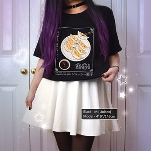 May include: Black t-shirt with a graphic of a plate of dumplings and chopsticks. The text on the shirt says "I'm so into you... I'll never let you go... Juicy Gyoza!" and "定番!" in Japanese. The model is wearing a white skirt and is 4' 9" tall.