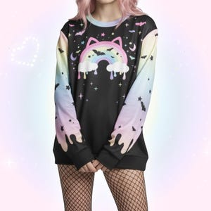 Puede incluir: Sudadera negra de manga larga con un diseño de gato arcoíris. Las mangas se desvanecen de azul a amarillo, con murciélagos y estrellas. El diseño incluye un arcoíris, nubes y un efecto de pintura goteando.