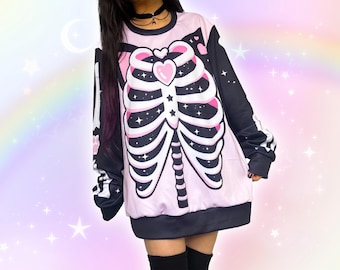 May include: A light pink sweatshirt with a black and white skeleton rib cage design. The long-sleeved top features a heart-shaped design on the chest and a black hemline. The sleeves are black with white bone details. The background is a pastel rainbow with stars.