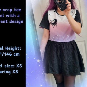 May include: A cropped t-shirt with a white background and a pink to purple ombre design. The shirt has a black crescent moon with a dripping black design and white stars. The model is wearing a black skirt and is 4'9"/146 cm tall.  Model size: XS. Wearing XS.