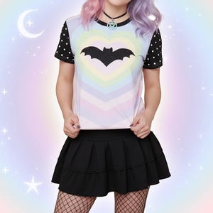 May include: A black bat graphic is centered on a pastel rainbow heart design on a short-sleeved t-shirt. The shirt has black sleeves with white polka dots. A black pleated skirt and fishnet stockings complete the outfit.