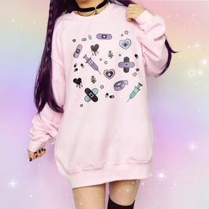 Könnte beinhalten: Rosafarbener Pullover mit einem grafischen Druck verschiedener medizinischer Symbole, darunter Verbände, Herzen, Spritzen und Pillen. Der Druck ist in Rosa-, Lila- und Blautönen gehalten.