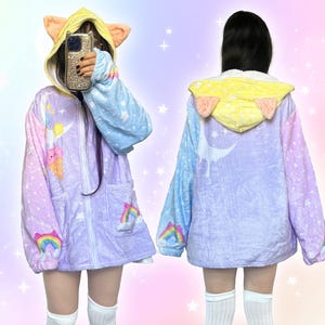 Puede incluir: Una bata con capucha de color pastel con un diseño de orejas de gato. La bata presenta cierre de cremallera, bolsillos y un patrón caprichoso de arcoíris, estrellas y conos de helado. Las mangas son azules y la capucha es amarilla.