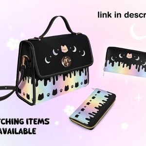 May include: Black satchel bag with a rainbow drip design and a cat face with crescent moons. The bag has a gold clasp and a matching wallet with the same design.
